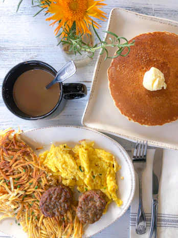 A breakfast setting with a black mug of coffee, a stack of pancakes topped with butter, a plate of scrambled eggs, hash browns, and sausage patties. A fork and knife are placed beside a folded napkin on the table. Bright orange flowers in a small vase are visible in the background.