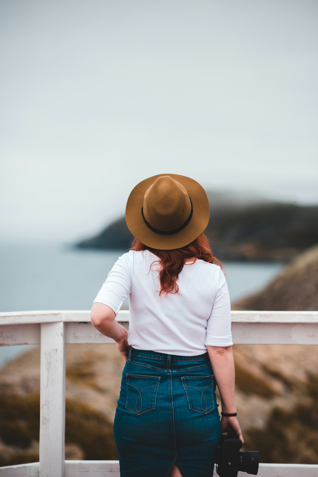 woman in white shirt and blue denim shorts wearing brown hat standing near body of water