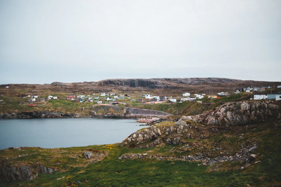 The image depicts a serene coastal landscape with a rocky cliff overlooking a calm body of water, possibly an ocean or sea. The shoreline is dotted with several small houses and buildings, suggesting a quaint village nestled on the hillside. The sky above is overcast, casting a soft light over the scene and creating a tranquil atmosphere.