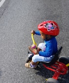A young child with a big smile riding a bright red adaptive tricycle on a sunny day in Oxford.