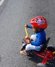 A child wearing a red helmet with a flower design rides a tricycle. The kid is dressed in a blue top, white pants, and brown shoes. The tricycle features a red and yellow frame and is being ridden on a paved road.