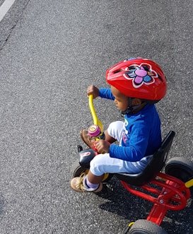A child wearing a red helmet with a flower design rides a tricycle. The kid is dressed in a blue top, white pants, and brown shoes. The tricycle features a red and yellow frame and is being ridden on a paved road.