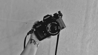 A monochrome photo featuring a hand holding a vintage SLR camera against a plain, textured background. The camera strap hangs loosely, and there is a tattoo visible on the wrist of the hand.