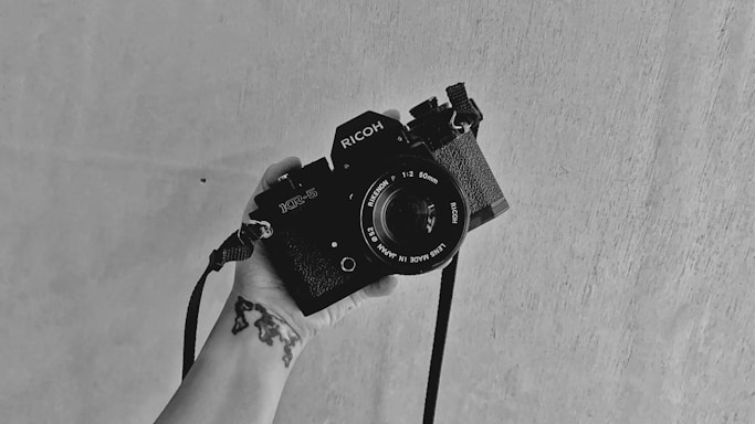 A monochrome photo featuring a hand holding a vintage SLR camera against a plain, textured background. The camera strap hangs loosely, and there is a tattoo visible on the wrist of the hand.