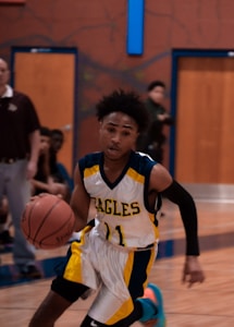 A basketball player in a white and yellow 'Eagles' jersey is dribbling a basketball on an indoor court. The player's number is 11, and he appears focused on the game. Other players and a referee are visible in the blurred background.