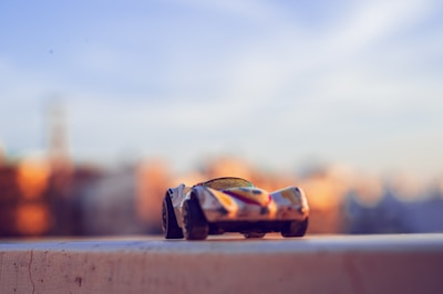 A small toy car is placed on a surface against a blurred background with soft lighting. The car is painted in multiple colors and appears to be a racing car model.
