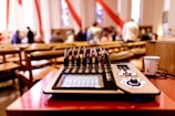 A sound mixer is placed on a red table in a room with wooden chairs and blurred people in the background, suggesting a gathering or event. Red and white drapes hang from the ceiling, and stained glass windows add colorful patterns to the walls.