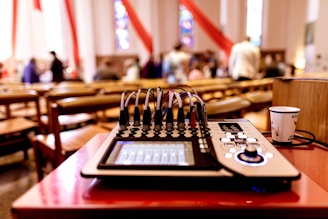 A sound mixer is placed on a red table in a room with wooden chairs and blurred people in the background, suggesting a gathering or event. Red and white drapes hang from the ceiling, and stained glass windows add colorful patterns to the walls.