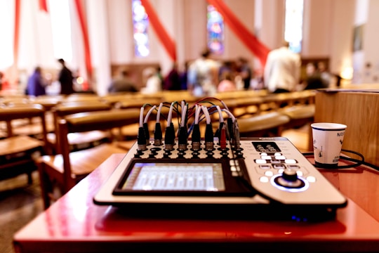 A sound mixer is placed on a red table in a room with wooden chairs and blurred people in the background, suggesting a gathering or event. Red and white drapes hang from the ceiling, and stained glass windows add colorful patterns to the walls.
