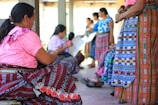 Women wearing traditional, brightly colored clothing, gathered together. The scene includes intricate patterns and vibrant textiles. They appear to be engaged in an activity, possibly weaving, within a sheltered outdoor area.