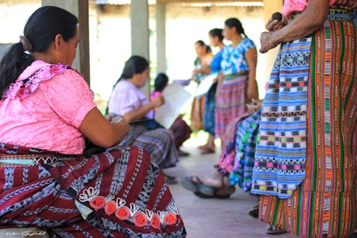 Women wearing traditional, brightly colored clothing, gathered together. The scene includes intricate patterns and vibrant textiles. They appear to be engaged in an activity, possibly weaving, within a sheltered outdoor area.