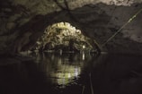 A natural cave with a dark, textured ceiling and walls, reflecting on the still water below. Light filters through an opening, illuminating the stalactites and rock formations inside. A metal railing can be seen, suggesting a defined path or walkway.