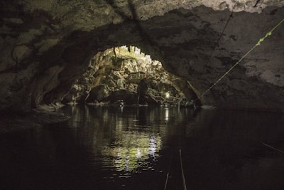 A natural cave with a dark, textured ceiling and walls, reflecting on the still water below. Light filters through an opening, illuminating the stalactites and rock formations inside. A metal railing can be seen, suggesting a defined path or walkway.