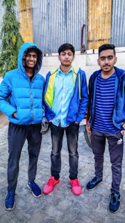 Three young men stand closely together, each wearing casual attire with jackets. The one on the left is smiling and wearing a bright blue hooded jacket, the middle individual has a serious expression and sports a blue and yellow jacket with bright red shoes, and the person on the right is dressed in a striped blue shirt with a dark blue jacket. The setting appears to be outdoors with a corrugated metal wall and some greenery in the background.