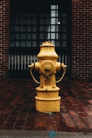 A vibrant yellow fire hydrant stands prominently against a backdrop of a brick wall and wet, red-colored brick pavement. The fireplace is surrounded by a rusted chain, and the lighting is low, giving a moody atmosphere to the scene.