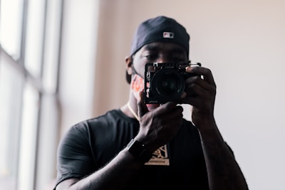 A person wearing a cap holds a camera up to their face. The background is softly lit, with a window to the left letting light into the room.