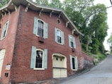A historic, two-story brick building features arched windows with pale green shutters and decorative corbels under the eaves. The structure is set on a sloped street surrounded by lush green trees. Its rustic charm is highlighted by small architectural details and a sign mounted near the corner.