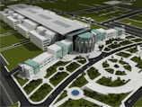 An aerial view of a modern architectural complex featuring a large central building with a circular glass facade and multiple adjoining rectangular structures. The complex is surrounded by landscaped gardens, pathways, and circular water features. A road with cars encircles the area, and parking spaces are visible in the foreground.