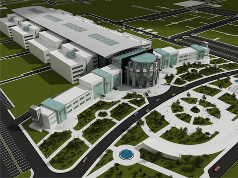 An aerial view of a modern architectural complex featuring a large central building with a circular glass facade and multiple adjoining rectangular structures. The complex is surrounded by landscaped gardens, pathways, and circular water features. A road with cars encircles the area, and parking spaces are visible in the foreground.