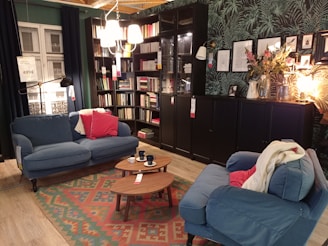 A cozy living room setup with two blue sofas accented by red and beige cushions. There is a wooden coffee table with several dark cups on it over a colorful patterned rug. The wall features a large bookshelf filled with books next to a dark cabinet with decorative items on top, including framed pictures and a plant. A unique light fixture hangs above, and a lamp provides additional warm lighting.