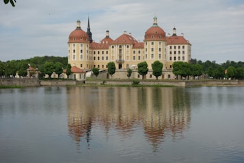 A large, elegant baroque-style castle with red domes and white and beige walls sits majestically by a tranquil lake, which reflects its beautiful architecture. The castle is surrounded by lush greenery, with neatly trimmed trees and a stone embankment along the water's edge.