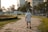 An elderly man is walking alone along a stone-paved path in a park. Surrounding him are green lawns and neatly trimmed bushes, with trees scattered throughout the area. The background shows urban buildings and a few parked cars, suggesting a city park setting. The lighting suggests it may be late afternoon or early evening.
