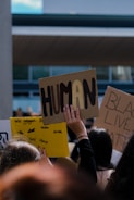 A group of people in a gathering hold various protest signs. The most prominent sign in the foreground reads 'HUMAN' in large, bold letters. Another sign in the background features the words 'BLACK LIVES MATTER'. The scene takes place outdoors with a modern building in the background.