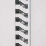 A minimalist design featuring the side view of a modern building with a series of white staircases zigzagging upwards against a plain white wall.