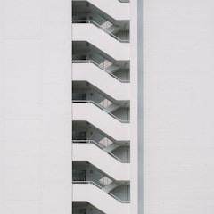A minimalist design featuring the side view of a modern building with a series of white staircases zigzagging upwards against a plain white wall.