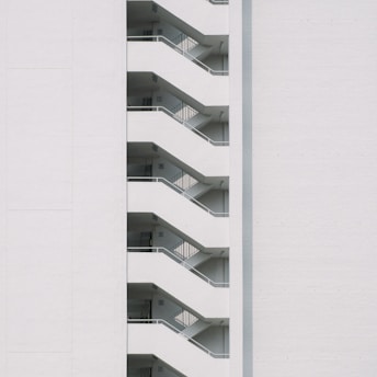 A minimalist design featuring the side view of a modern building with a series of white staircases zigzagging upwards against a plain white wall.