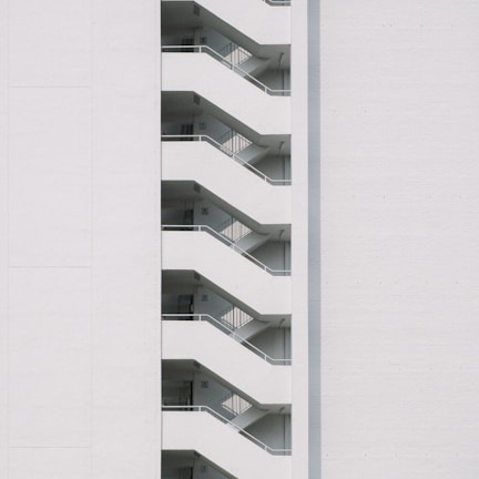 A minimalist design featuring the side view of a modern building with a series of white staircases zigzagging upwards against a plain white wall.