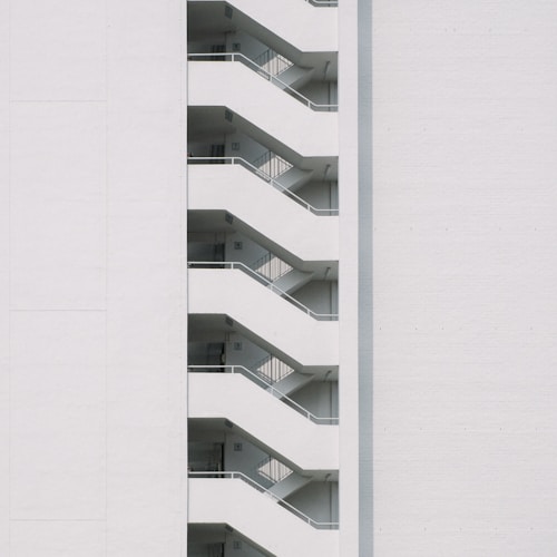 A minimalist design featuring the side view of a modern building with a series of white staircases zigzagging upwards against a plain white wall.