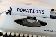 A vintage typewriter with a sheet of paper displaying the word 'DONATIONS' in bold uppercase letters. The typewriter is on a wooden surface with a soft focus on the background.