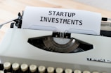 A vintage typewriter with a piece of paper displaying the text 'STARTUP INVESTMENTS'. The typewriter is a classic model with keys in focus and the paper neatly inserted into the ribbon area.