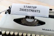A vintage typewriter with a piece of paper displaying the text 'STARTUP INVESTMENTS'. The typewriter is a classic model with keys in focus and the paper neatly inserted into the ribbon area.