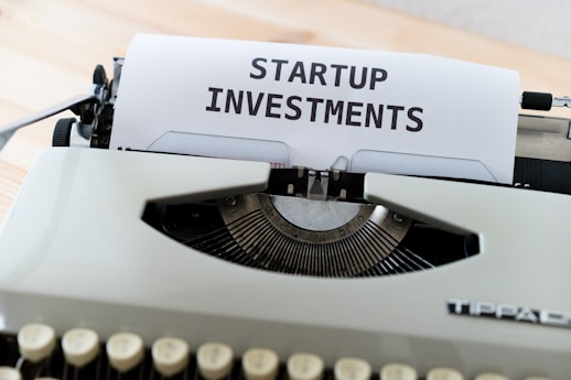 A vintage typewriter with a piece of paper displaying the text 'STARTUP INVESTMENTS'. The typewriter is a classic model with keys in focus and the paper neatly inserted into the ribbon area.