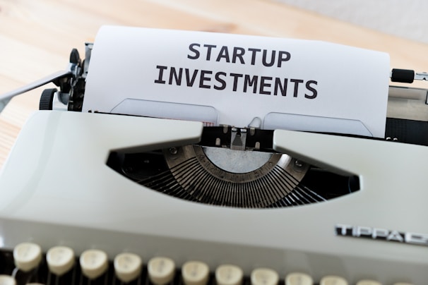 A vintage typewriter with a piece of paper displaying the text 'STARTUP INVESTMENTS'. The typewriter is a classic model with keys in focus and the paper neatly inserted into the ribbon area.