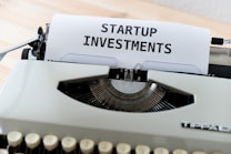 A vintage typewriter with a piece of paper displaying the text 'STARTUP INVESTMENTS'. The typewriter is a classic model with keys in focus and the paper neatly inserted into the ribbon area.