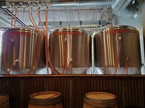 Three large stainless steel fermentation vessels are aligned in a row inside a brewery. Each vessel is connected to pipes and hoses, with inscriptions indicating their numbering: #3, #2, and #1 from left to right. The setting includes industrial elements such as metal pipes and ceiling fixtures, suggesting a functional brewing space. Wooden barrels are also visible at the forefront, contributing to the ambiance.