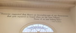 A wall features a quote painted in black text: 'Someone suggested that there's an incomplete part of our chromosomes that gets repaired or found when we hit New Orleans. Some of us just belong here.' Attributed to John Goodman, the quote is displayed beside a semi-circular window that reveals a glimpse of the outdoors.