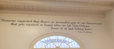 A wall features a quote painted in black text: 'Someone suggested that there's an incomplete part of our chromosomes that gets repaired or found when we hit New Orleans. Some of us just belong here.' Attributed to John Goodman, the quote is displayed beside a semi-circular window that reveals a glimpse of the outdoors.