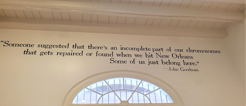 A wall features a quote painted in black text: 'Someone suggested that there's an incomplete part of our chromosomes that gets repaired or found when we hit New Orleans. Some of us just belong here.' Attributed to John Goodman, the quote is displayed beside a semi-circular window that reveals a glimpse of the outdoors.
