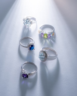 A collection of five elegant rings, each set with different gemstones, arranged on a soft blue surface. The rings feature diverse designs, including floral and geometric settings, with gemstones in shades of blue, purple, and green. Light shines from the top right, casting subtle reflections and shadows.