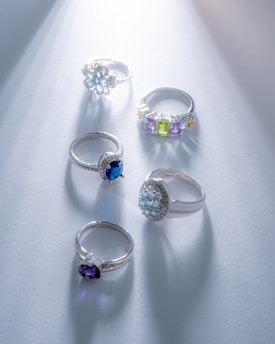 A collection of five elegant rings, each set with different gemstones, arranged on a soft blue surface. The rings feature diverse designs, including floral and geometric settings, with gemstones in shades of blue, purple, and green. Light shines from the top right, casting subtle reflections and shadows.