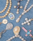 A collection of religious-themed jewelry arranged on a blue denim fabric. The pieces include beaded rosaries, pendants, and medals featuring pearls and gold accents, with some displaying religious icons and symbols.