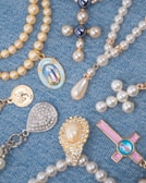 A collection of religious-themed jewelry arranged on a blue denim fabric. The pieces include beaded rosaries, pendants, and medals featuring pearls and gold accents, with some displaying religious icons and symbols.