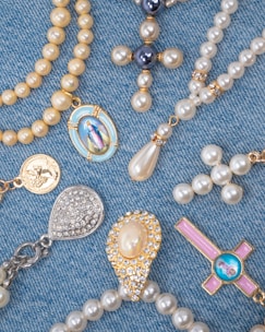 A collection of religious-themed jewelry arranged on a blue denim fabric. The pieces include beaded rosaries, pendants, and medals featuring pearls and gold accents, with some displaying religious icons and symbols.
