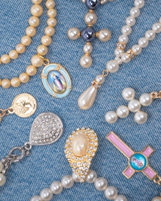 A collection of religious-themed jewelry arranged on a blue denim fabric. The pieces include beaded rosaries, pendants, and medals featuring pearls and gold accents, with some displaying religious icons and symbols.