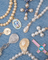 A collection of religious-themed jewelry arranged on a blue denim fabric. The pieces include beaded rosaries, pendants, and medals featuring pearls and gold accents, with some displaying religious icons and symbols.