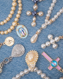 A collection of religious-themed jewelry arranged on a blue denim fabric. The pieces include beaded rosaries, pendants, and medals featuring pearls and gold accents, with some displaying religious icons and symbols.